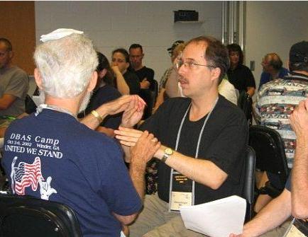 Ed Knight is serving as SSP for Steven Frank, a delegate to the AADB convention.  Steven is receiving in tactile as Ed signs,  shadowing the signs of the speaker, who is a deaf rabbi who is out of camera range.  Steven has a folded handkercheif on his head to serve as a substitute yarmulke.