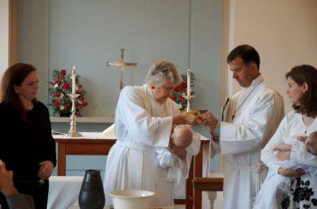 Two godparents watch as Mother Barbara receives chrism from LEM Tom Hattaway, holding baby in other arm.   Everyone seems serene including the twins.