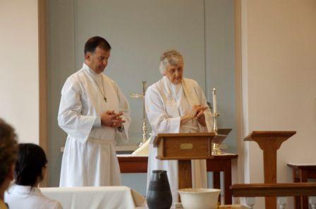 Tom Hattaway in alb and Mother Barbara in white stole and altar linens in white, for the baptism.  In foreground is ceramic pitcher with warm water and bowl for baptism.